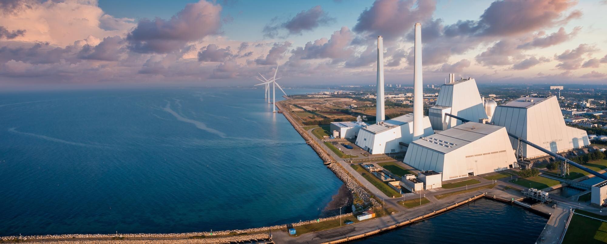 Aerial view of a coastal power station with twin chimneys, waterfront roads and offshore wind turbines.